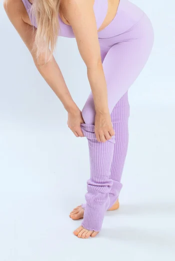 Lähivaade naisest, kes tõmbab jalga lavendli kootud joogasääristeid. Sääristel on avatud koha ja varbaosa disain ning need on kantud sarnast tooni lillade retuuside peal. Pilt rõhutab sääriste pehmet soonikkoes tekstuuri ja praktilisust.